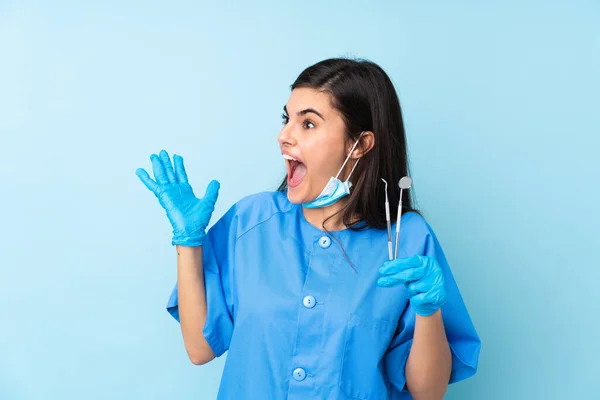 年轻的女牙医把工具放在孤立的蓝色背景上 满脸惊讶的表情 — 图库照片