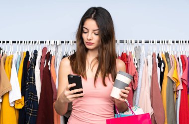 Kafkasyalı genç bir kadın marketten kıyafet alıyor elinde kahve ve cep telefonu var.