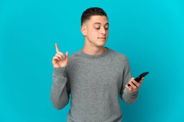 Genç beyaz adam mavi arka planda cep telefonu ve parmak kaldırarak izole edildi.