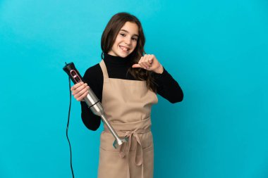 Mavi arka planda izole edilmiş el blenderi kullanan küçük kız gururlu ve kendini beğenmiş.