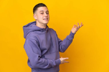 Sarı arka planda izole edilmiş genç beyaz adam davet etmek için ellerini yana uzatıyor.