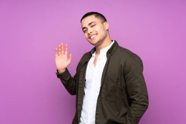 Asyalı yakışıklı adam mavi arka planda izole edilmiş, mutlu bir ifadeyle selamlıyor.