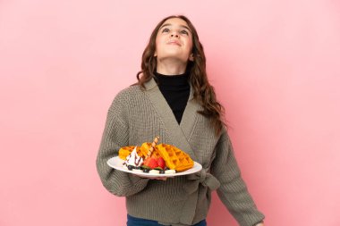 Elinde waffle tutan küçük kız pembe arka planda izole edilmiş ve yukarı bakıyor.