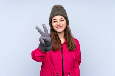 Kış şapkalı genç kız mavi arka planda izole edilmiş gülümsüyor ve zafer işareti gösteriyor.