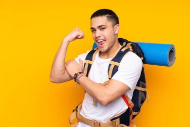 Sarı arka planda izole edilmiş büyük sırt çantalı Asyalı genç adam güçlü bir jest yapıyor.