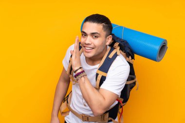 Sarı arka planda izole edilmiş büyük sırt çantalı Asyalı genç adam bir şeyler fısıldıyor.