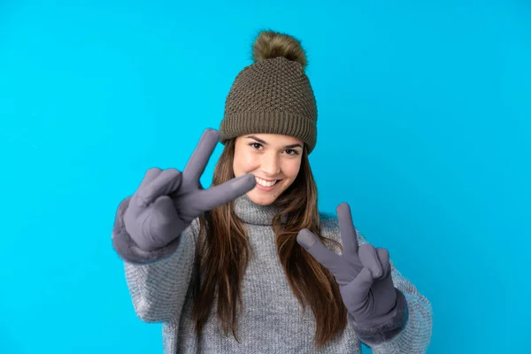 Kışlık Şapkalı Genç Kız Mavi Arka Planda Gülümsüyor Zafer Işareti — Stok fotoğraf