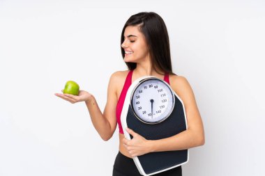 Soyutlanmış beyaz arka plan üzerinde genç bir kadın bir elma ararken bir tartı makinesi tutuyor.