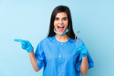 İzole edilmiş mavi arka planda aletleri tutan genç dişçi kadın şaşırdı ve parmağını yana çevirdi.