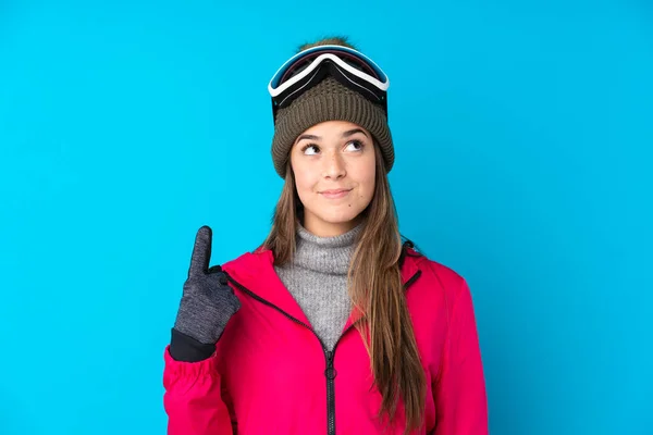 年轻的滑雪者女孩 戴着雪板眼镜 背景是蓝色的 她用食指指尖着一个很好的主意 — 图库照片