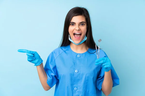 Młoda Kobieta Dentysta Trzyma Narzędzia Odizolowanym Niebieskim Tle Zaskoczony Wskazując — Zdjęcie stockowe