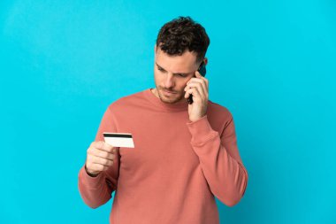 Kafkasyalı yakışıklı genç adam mavi arka planda izole edilmiş ve kredi kartıyla cep telefonuyla alışveriş yapıyor.