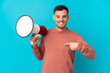 Mavi arka planda, elinde bir megafon ve yüzünde sürpriz bir ifadeyle izole edilmiş beyaz, yakışıklı bir genç.