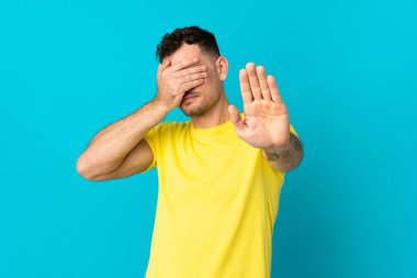 Beyaz tenli yakışıklı adam mavi arka planda izole edilmiş. Jesti durdurup yüzünü kapatıyor.