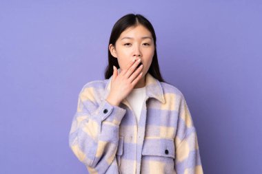 Mor arka planda izole edilmiş genç Asyalı kadın esniyor ve ağzını eliyle açık tutuyor.