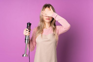 Genç Rus kız el blenderi kullanıyor. Mor arka planda izole edilmiş, gözleri elle kapatıyor. Bir şey görmek istemiyorum.