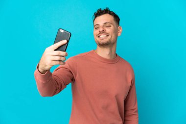 Kafkasyalı yakışıklı adam mavi arka planda izole edilmiş cep telefonuyla selfie çekiyor.