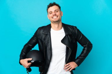 Motosiklet kasklı beyaz adam mavi arka planda izole edilmiş, kolları kalçasında ve gülümsüyor.