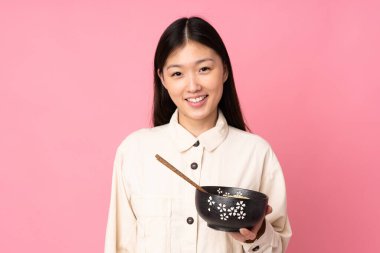 Pembe arka planda izole edilmiş genç Asyalı kadın elinde çubuklarla elinde bir kase erişte tutarken gülümsüyor.