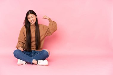 Genç Asyalı bir kadın pembe arka planda izole bir şekilde yerde oturmuş güçlü bir jest yapıyor.