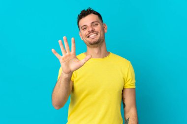 Beyaz tenli yakışıklı adam mavi arka planda izole edilmiş, parmaklarıyla beşi sayıyor.