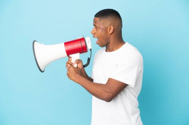 Genç latin adam mavi arka planda izole edilmiş. Megafonla bir şey duyurmak için bağırıyor.