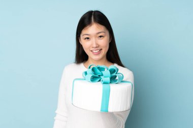 Asyalı pasta şefi elinde büyük bir pasta tutuyor mavi arka planda mutlu bir ifadeyle izole edilmiş.