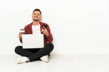 Genç, yakışıklı, beyaz bir adam dizüstü bilgisayarıyla yerde oturuyor. Elinde kahve ve cep telefonu var.