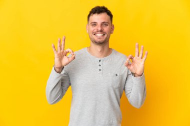 Sarı arka planda izole edilmiş beyaz, yakışıklı genç bir adam iki eliyle tamam işareti gösteriyor.