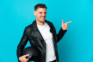 Motosiklet kasklı beyaz bir adam mavi arka planda izole edilmiş bir şekilde parmak ucunda bir ürün sunuyor.