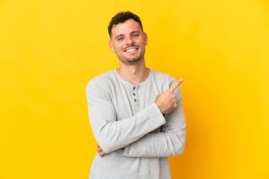 Sarı arka planda izole edilmiş beyaz, yakışıklı genç bir adam bir ürün sunmak için kenarı işaret ediyor.