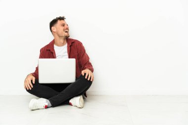 Genç, yakışıklı, beyaz bir adam dizüstü bilgisayarla yerde oturuyor ve yan pozisyonda gülüyor.