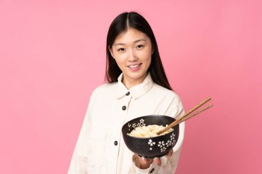 Pembe arka planda izole edilmiş genç Asyalı kadın elinde çubuklu erişte kasesi tutarken mutlu bir ifadeyle.