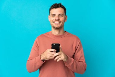 Beyaz tenli yakışıklı adam mavi arka planda izole edilmiş kameraya bakıyor ve cep telefonunu kullanarak gülümsüyor.