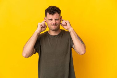 Sarı arka planda izole edilmiş genç beyaz yakışıklı adam hayal kırıklığına uğramış ve kulaklarını kapatmış.