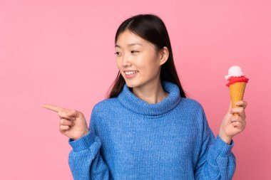 Kornet dondurması olan genç Asyalı bir kadın pembe arka planda izole edilmiş bir şekilde bir ürünü sunmak için kenarı işaret ediyor.
