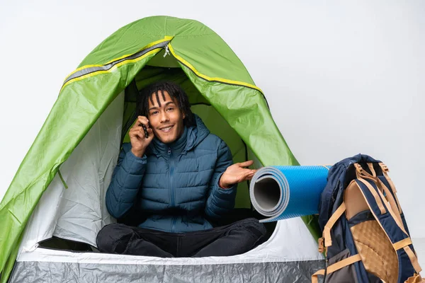 Young african american man inside a camping green tent keeping a conversation with the mobile phone with someone