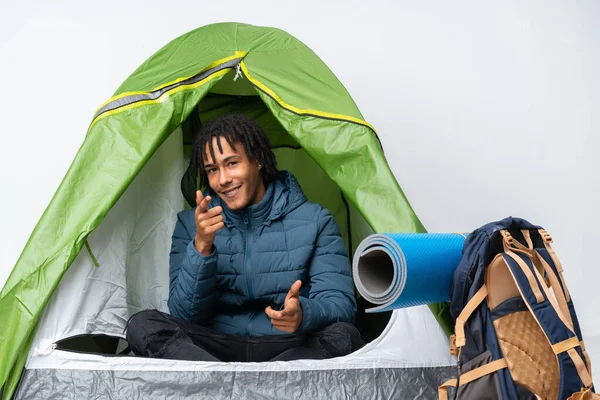 Young african american man inside a camping green tent pointing to the front and smiling