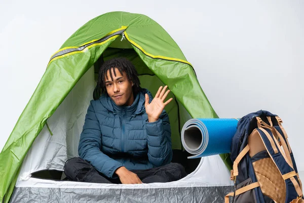 Young african american man inside a camping green tent saluting with hand with happy expression