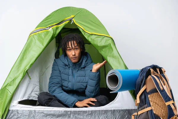 Young african american man inside a camping green tent making doubts gesture