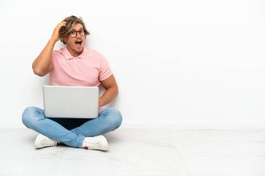 Beyaz arka planda dizüstü bilgisayarıyla yerde oturan genç beyaz bir adam yan tarafa bakarken sürpriz jest yapıyor.