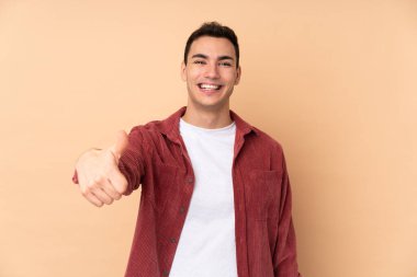 Beyaz tenli yakışıklı adam bej arka planda izole edilmiş. Başparmakları havada. Çünkü iyi bir şey oldu.