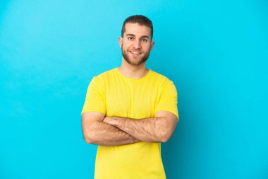 Mavi arka planda izole edilmiş yakışıklı beyaz adam kollarını ön cephede çapraz tutuyor.