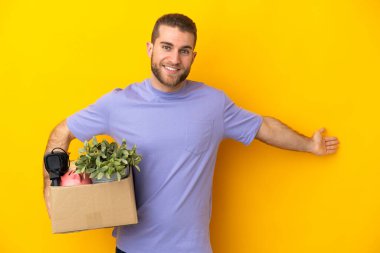 Genç Kafkasyalı, sarı arka planda izole edilmiş eşyalarla dolu bir kutuyu kaldırırken hareket ediyor. Davet etmek için ellerini yana açıyor.