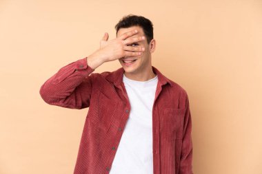 Beyaz tenli yakışıklı adam bej arka planda izole edilmiş gözlerini elleriyle kapatmış ve gülümsüyor.