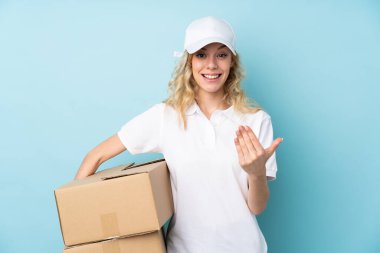 Genç teslimatçı kadın mavi arka planda izole edilmiş, davet ediliyor.