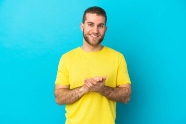 Mavi arka planda izole edilmiş yakışıklı beyaz adam alkışlıyor.