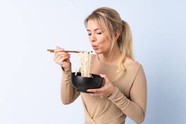 Sarışın kadın izole edilmiş mavi arka planda elinde çubuklu bir kase erişteyle sıcak olduğu için üflüyor.