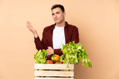 Bej arka planda izole edilmiş bir kutuda taze sebze toplayan çiftçi bir şeyi anlamadığı için mutsuz.