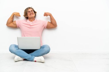 Genç, beyaz arka planda laptopuyla yerde oturan beyaz bir adam güçlü bir jest yapıyor.